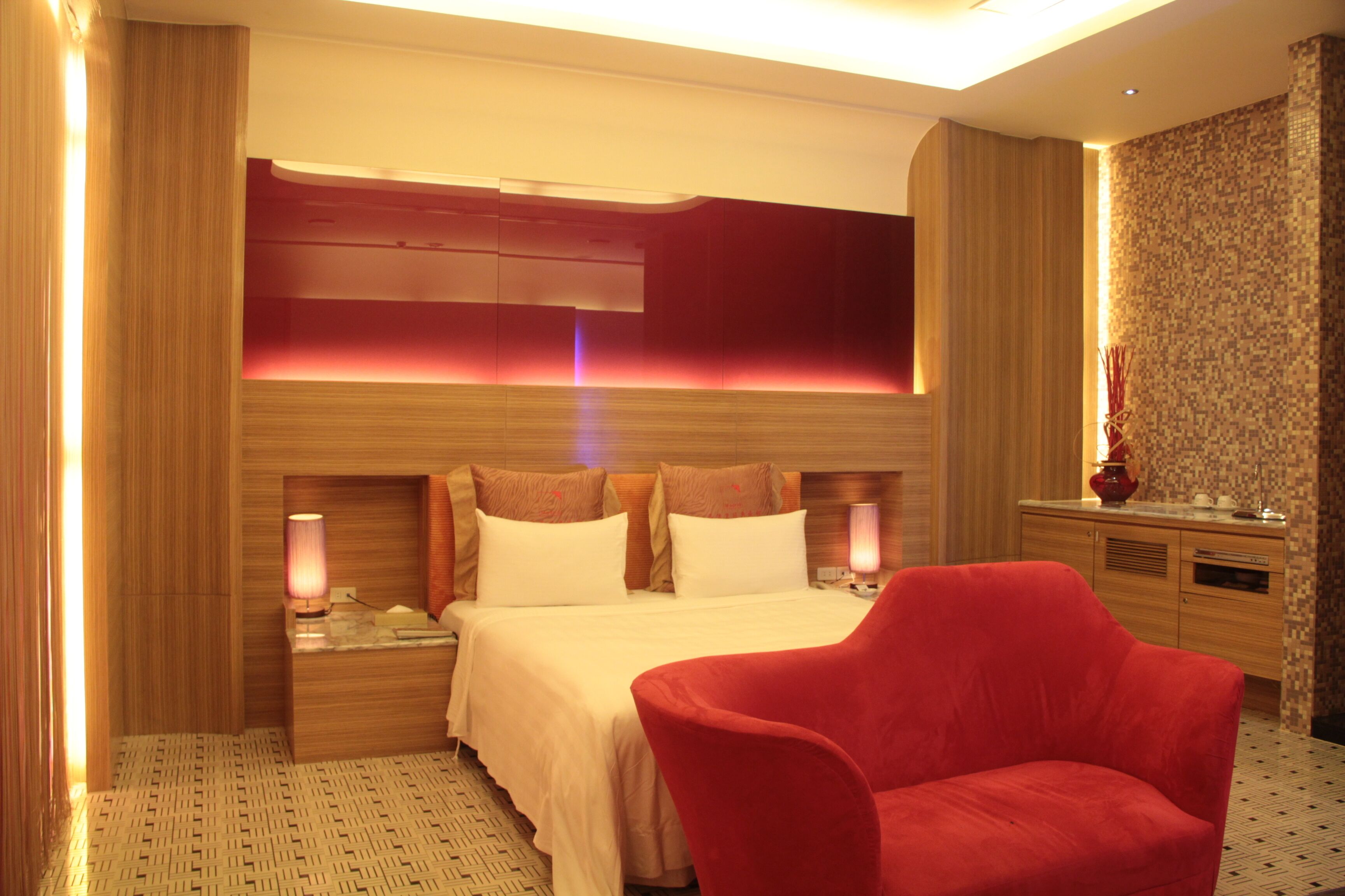 Une chambre d’hôtel avec un grand lit, deux tables de chevet avec des lampes, un fauteuil rouge et une tête de lit en bois.