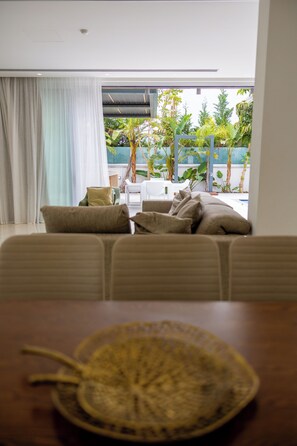 A living room with a sofa, a dining table with a placemat, and a view of a garden through a window.
