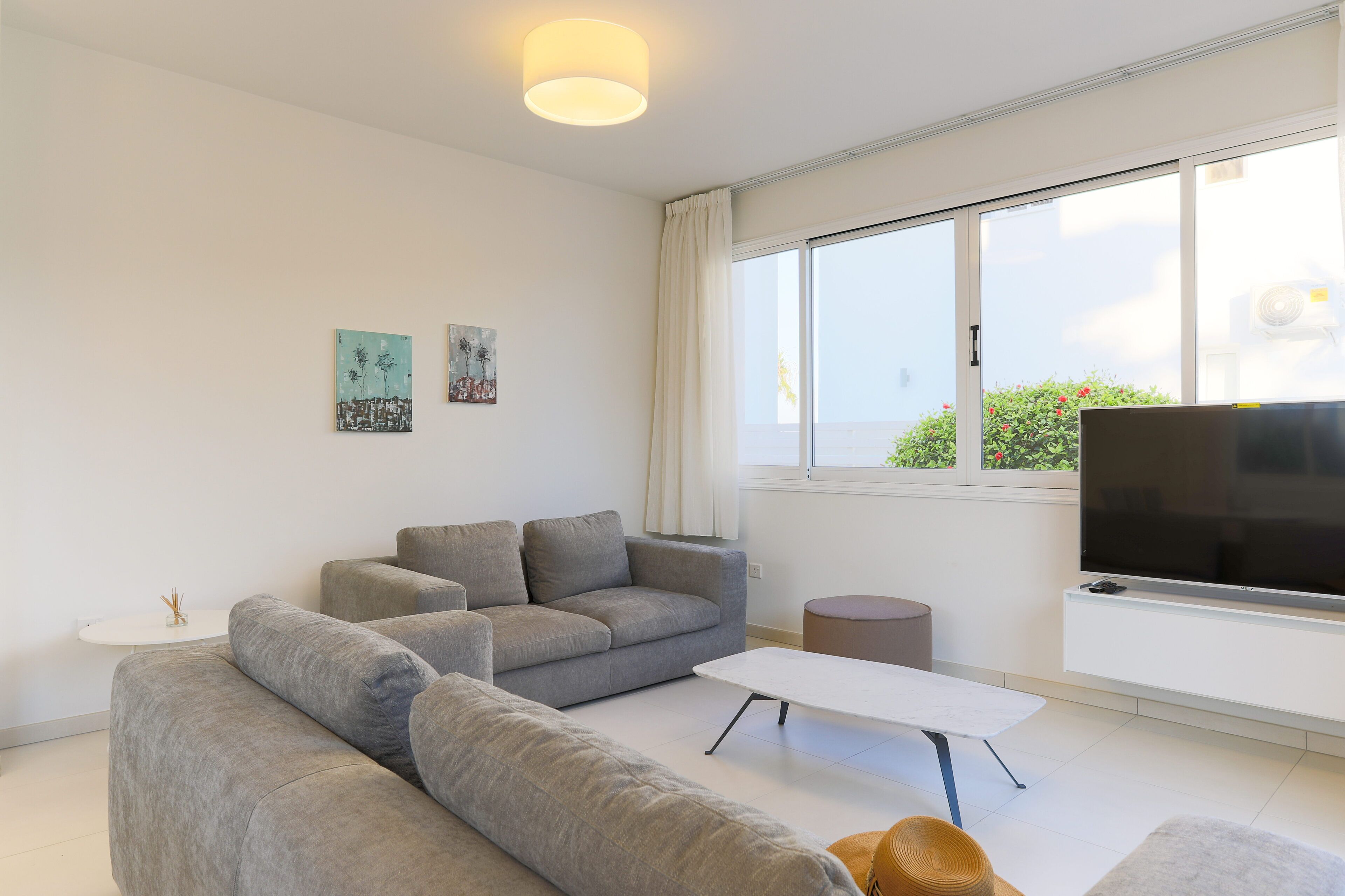 Een moderne woonkamer met een grijze bank, een witte salontafel en een flatscreen televisie aan de muur. Er hangen twee schilderijen aan de wand, en een raam met gordijnen.