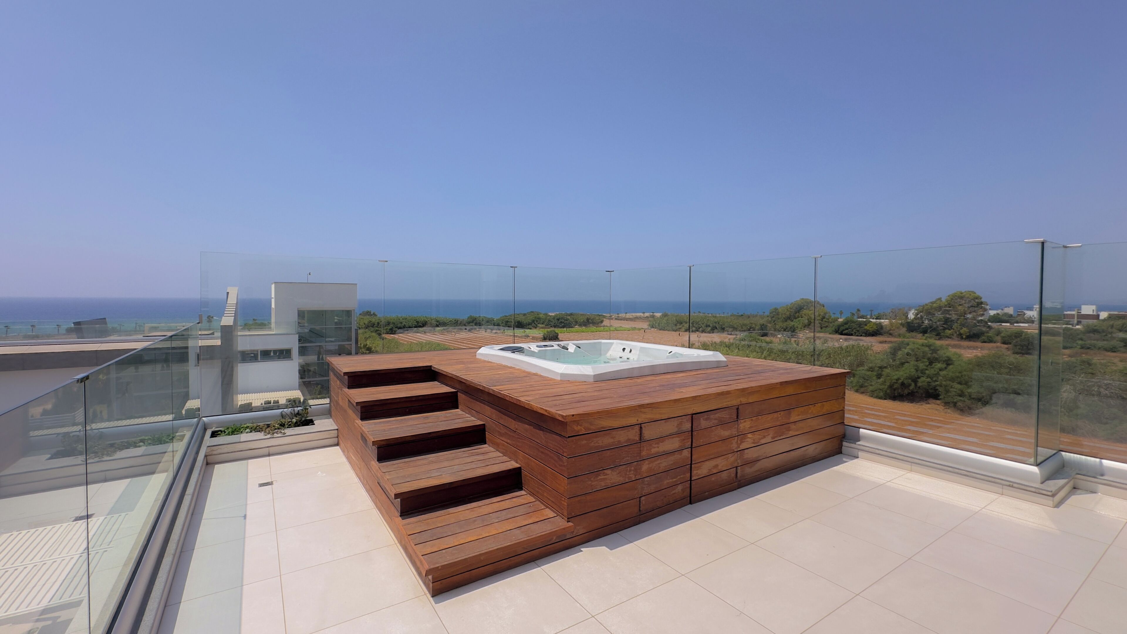 A rooftop terrace with a hot tub, wooden steps, and a glass railing overlooking a coastal landscape.