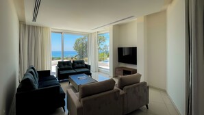 A modern living room with a sofa, armchairs, a coffee table, and a TV. Large windows offer a view of the ocean.