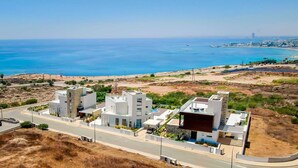 A modern building complex with multiple structures, a road in front, and a view of the sea and distant cityscape.