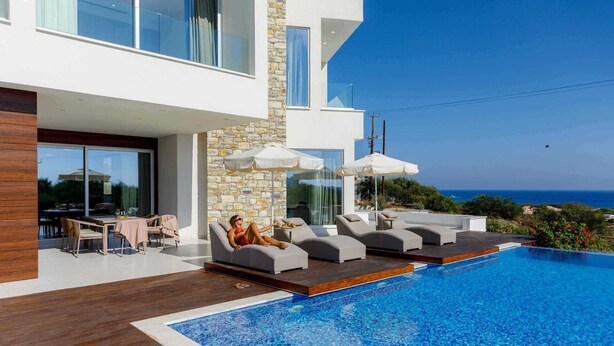 A modern outdoor pool area with lounge chairs and umbrellas, a dining area with a table and chairs, and a view of the sea.