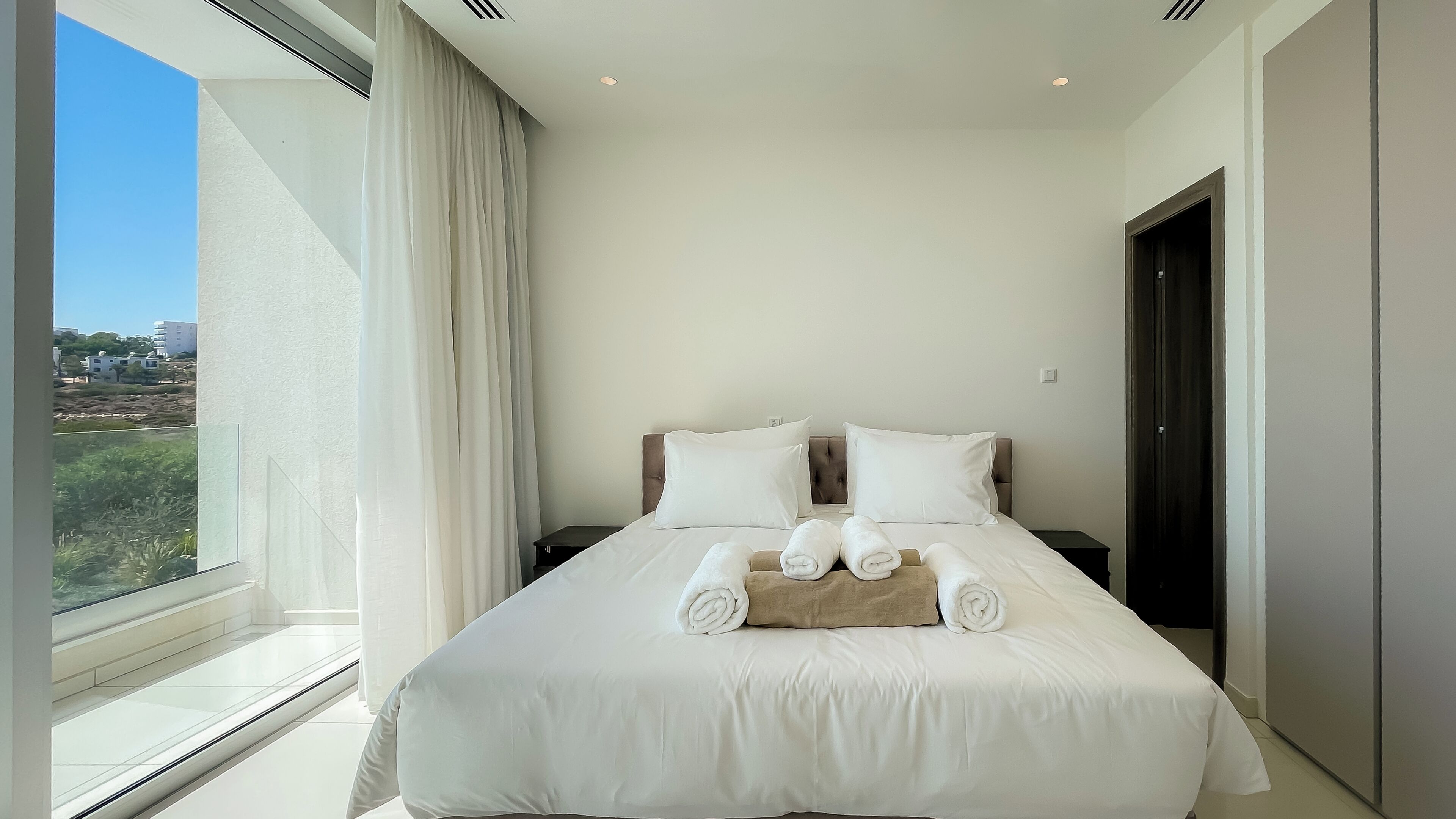 A hotel room with a large bed, two pillows, a nightstand, and a view of the outdoors.