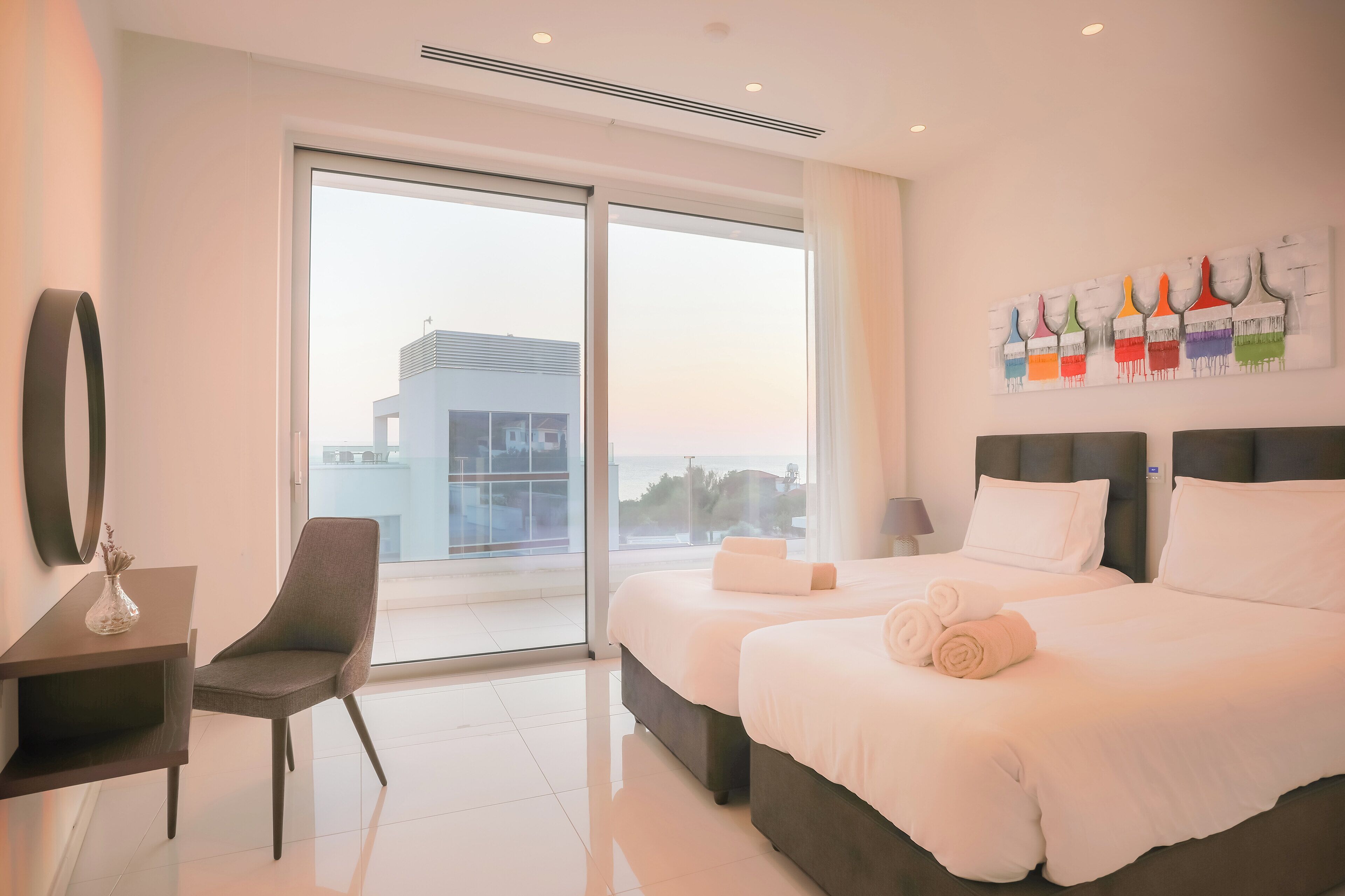 A modern hotel room with a large window, a desk with a chair, two beds with white linens, and a colorful abstract painting on the wall.