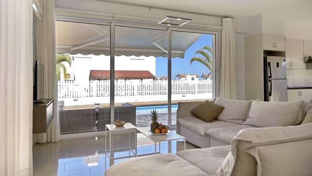 A modern living room with a white sofa, a small table with fruit, and a view of a pool and buildings. - Μake you feel at home. Quite location deal for Family (Paralimni)