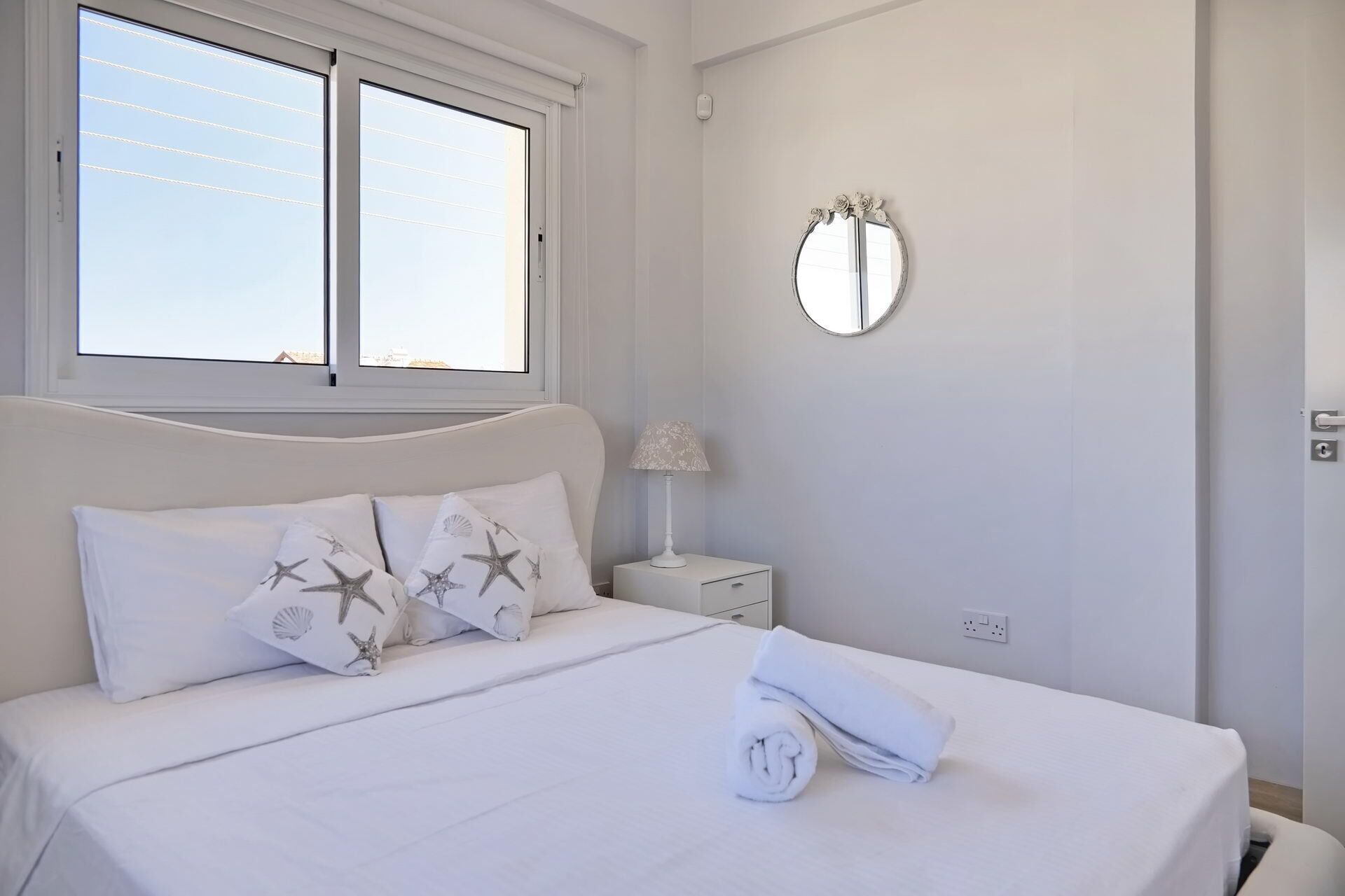 Un lit bien fait, avec une literie blanche et des oreillers à motifs étoilés, une lampe de chevet et un miroir rond accroché au mur.