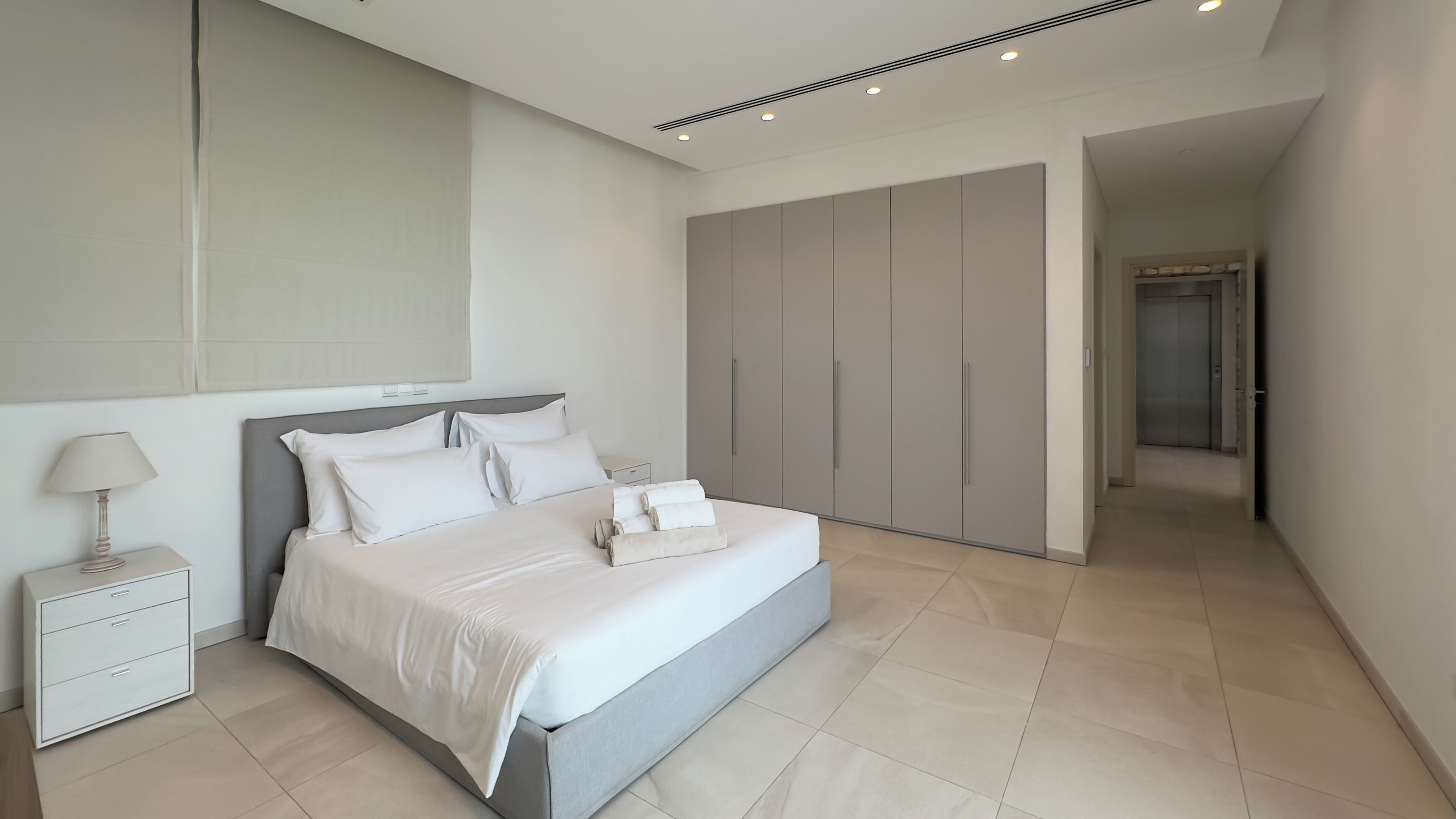 A modern bedroom with a large bed, a nightstand, a wardrobe, and a hallway leading to a door.