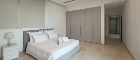 A modern bedroom with a large bed, a nightstand, a wardrobe, and a hallway leading to a door.