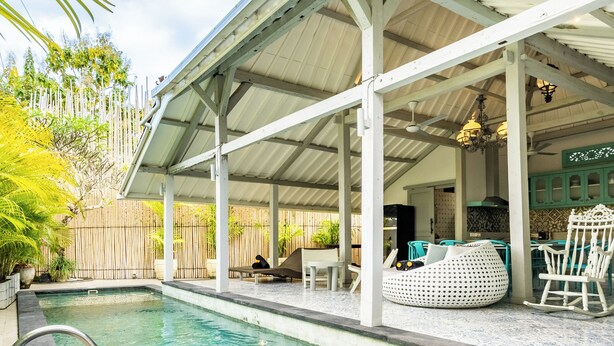Ein klares Schwimmbecken mit einer Metallleiter, umgeben von einer überdachten Terrasse mit Sitzgelegenheiten und einem Holzzaun.