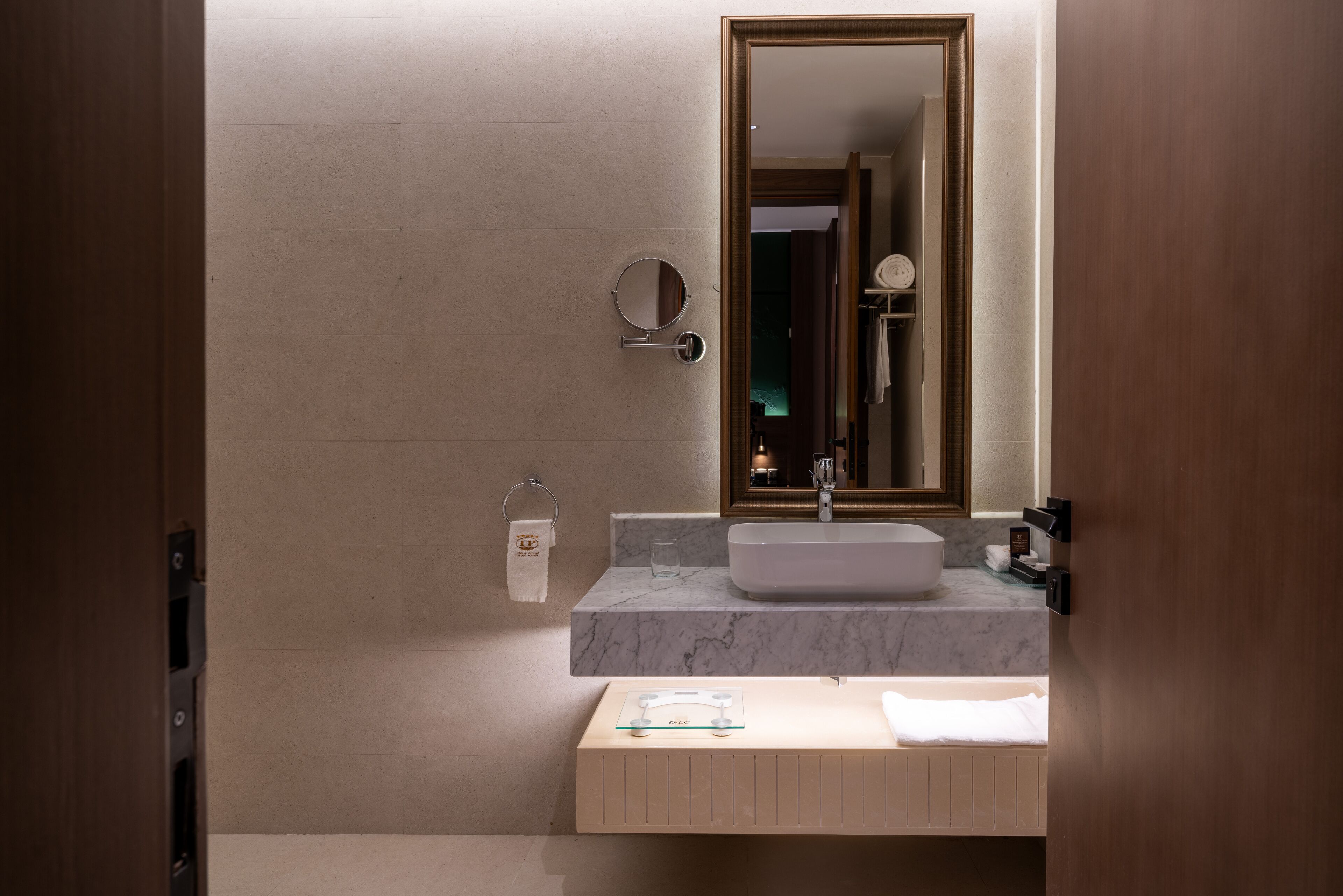 A modern bathroom with a marble countertop, a round mirror, a towel rack, and a wooden vanity.