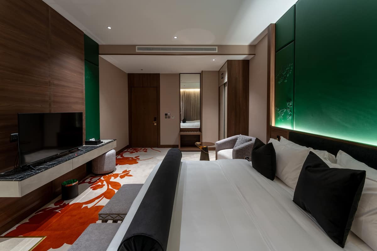 A modern hotel room with a flat-screen TV, a sofa, a bed with white and black pillows, and a green accent wall.