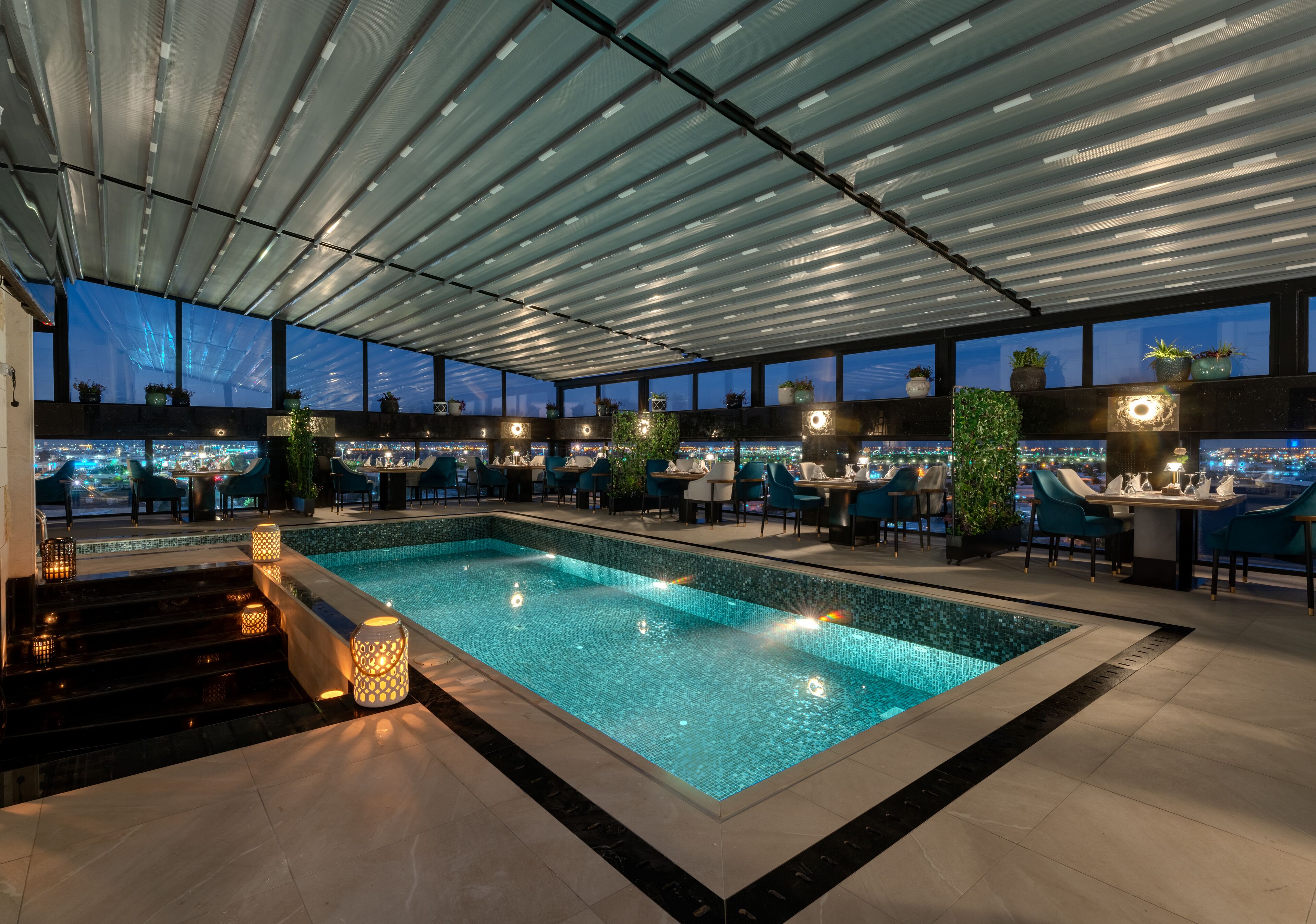 Espaço moderno de piscina interna com vista para a cidade à noite, rodeado por assentos e plantas.