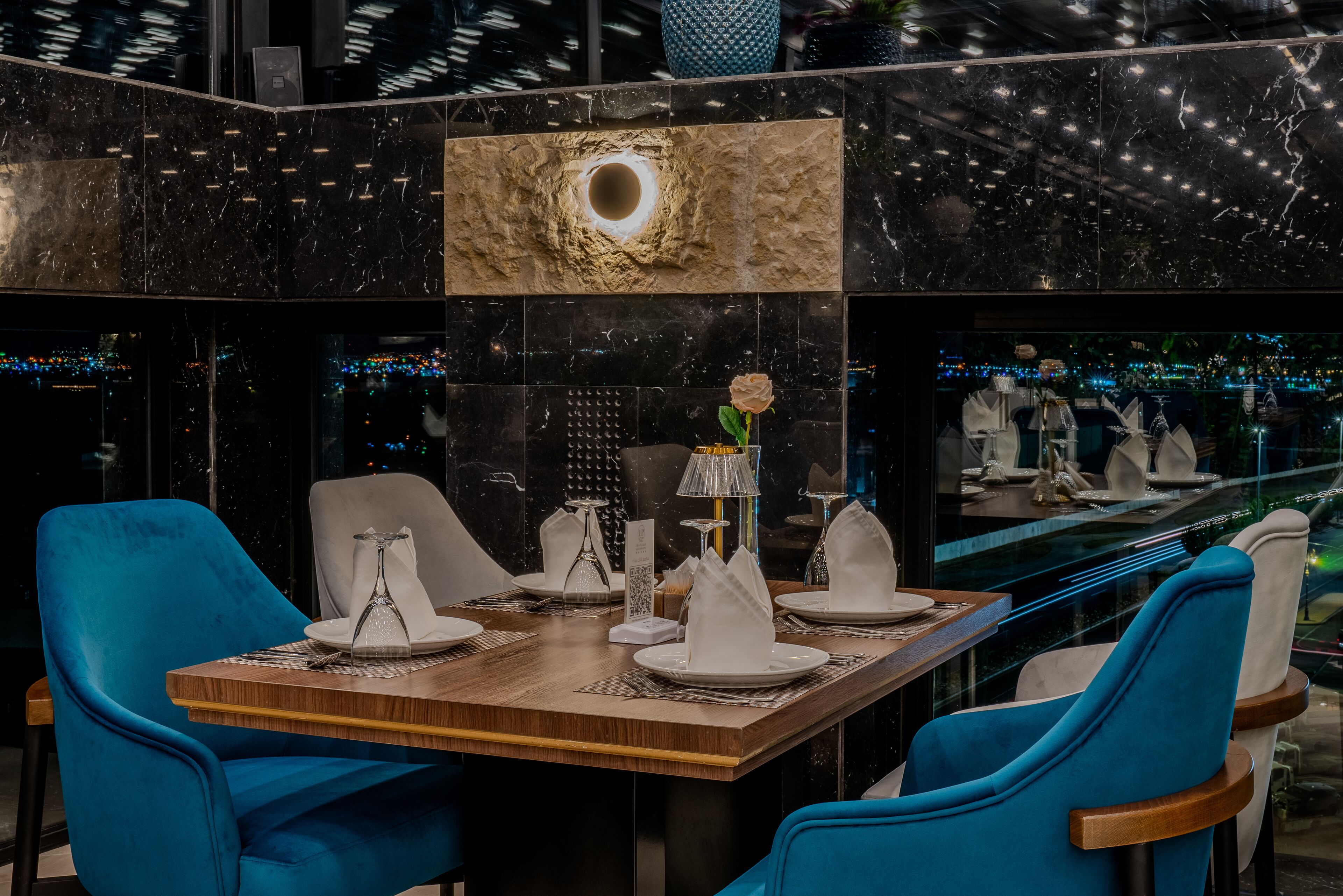 Un comedor moderno con una mesa de madera dispuesta para cuatro personas, sillas tapizadas en azul y un centro de mesa decorativo con rosas.