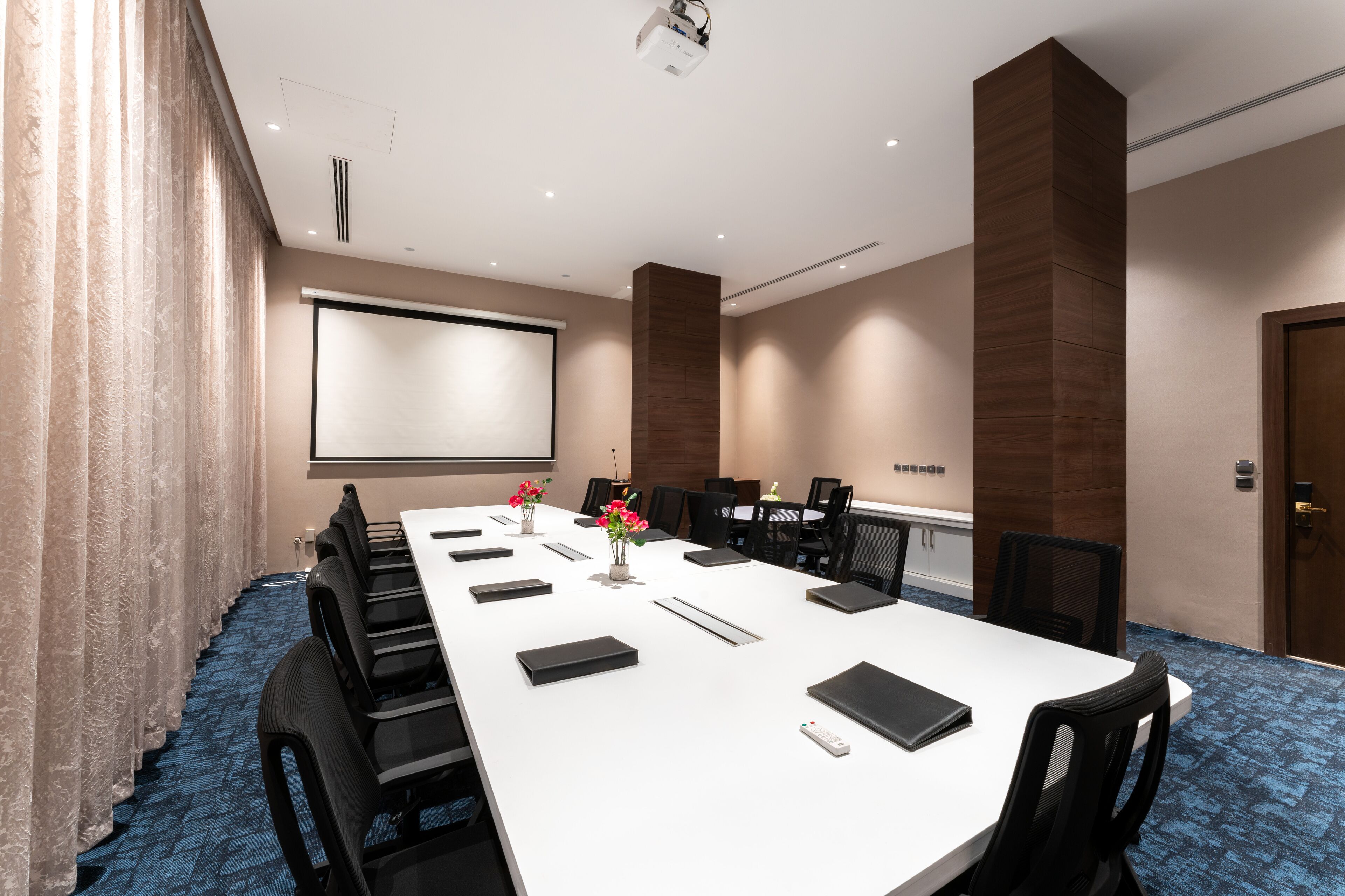 A conference room with a long table, chairs, a projector screen, and a blue carpet.