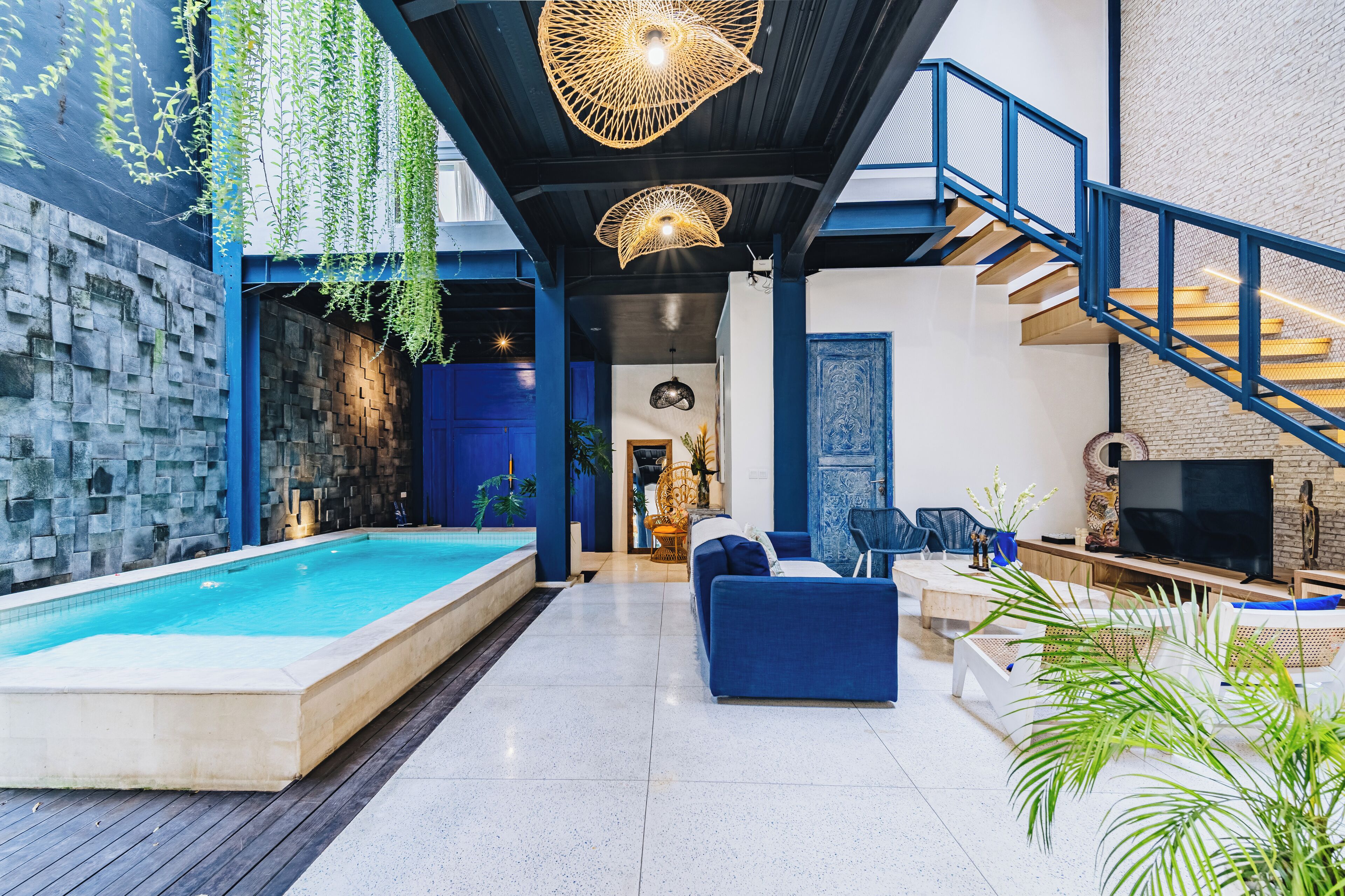 Un área de piscina interior moderna con un sofá azul, un televisor y plantas colgantes.