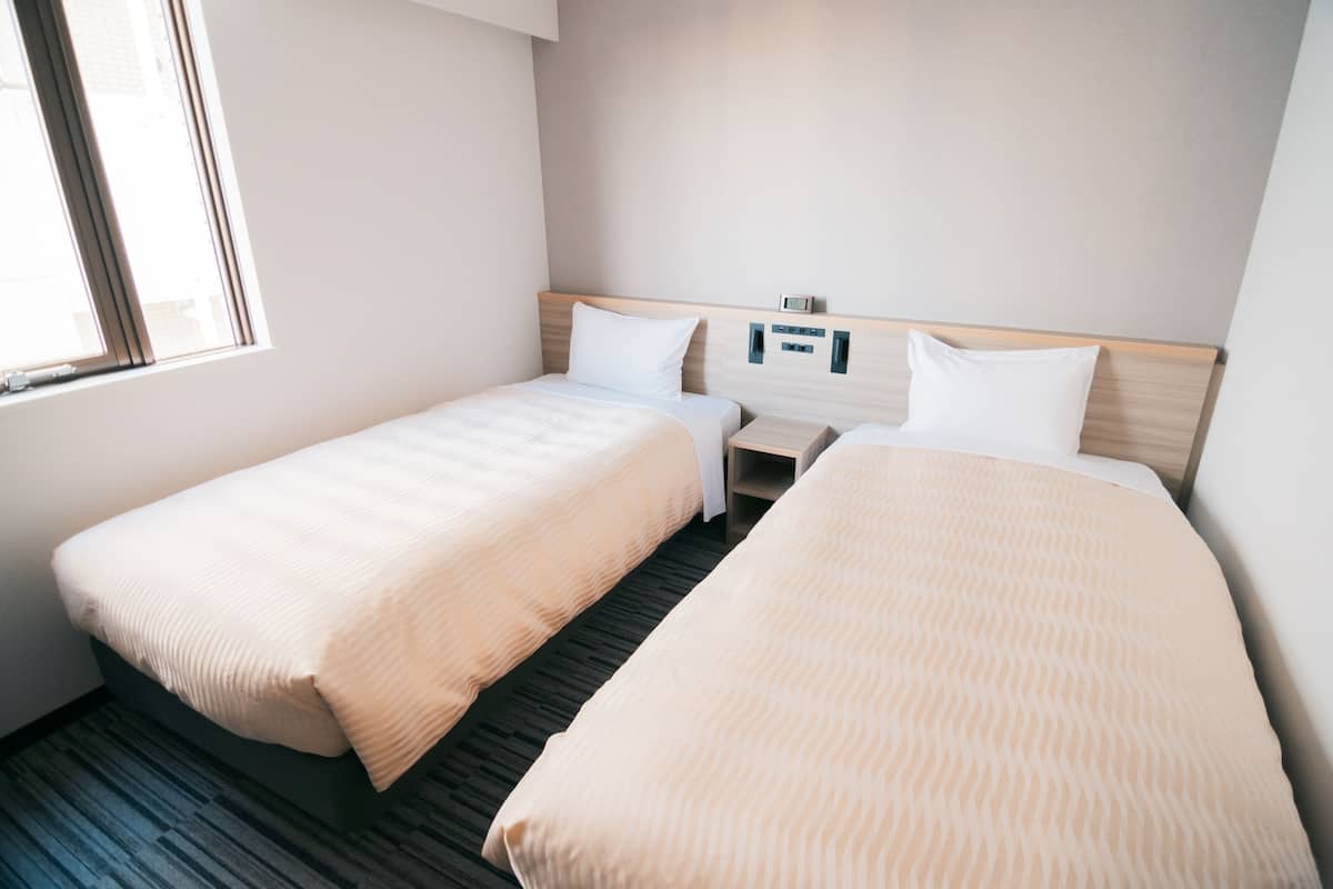 A hotel room with two single beds, a bedside table, a nightstand, and a window.