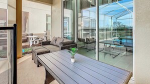 A sunlit patio with a dining table, cushioned seating, and a glass enclosure overlooking a swimming pool.