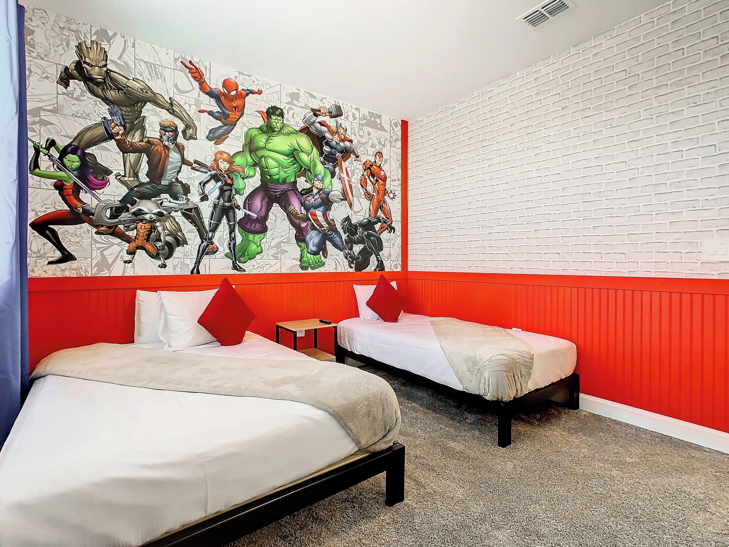 Une chambre d’hôtel avec deux lits, un mur d’accent rouge et une grande fresque murale sur le thème des super-héros.