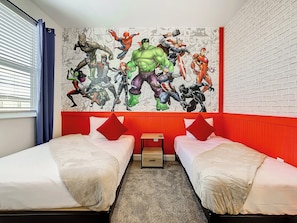 Quarto de hotel com duas camas, parede com detalhes em vermelho e um grande mural com tema de super-heróis.