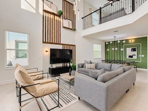 A spacious living area with a high ceiling, a flat-screen TV, a grey sofa, a beige armchair, and a dining table.