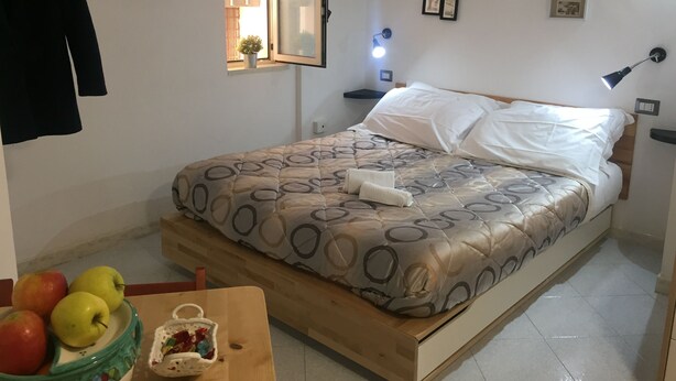 Um quarto com cama, mesa de cabeceira, abajur, janela e uma parede com quadros.