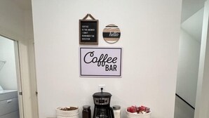 Un coin café aménagé avec une machine à café, des bocaux et des tasses sur une table en bois, le tout adossé à un mur blanc.