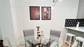 Un espace de salle à manger moderne avec une table en verre entourée de quatre chaises grises à revêtement. Deux affiches encadrées sont accrochées au mur au-dessus de la table, et un luminaire suspendu est fixé au plafond.