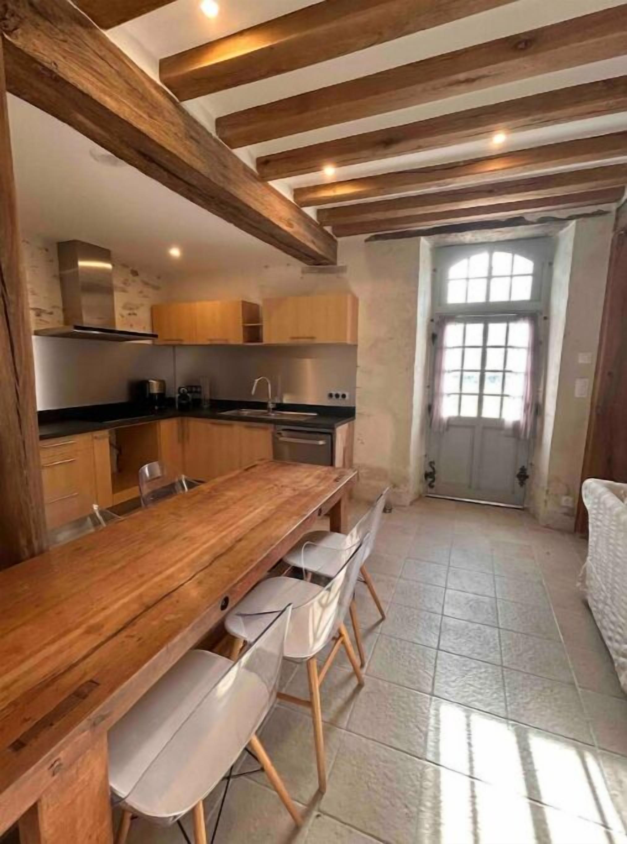 Une cuisine avec des poutres en bois, une longue table en bois et des chaises blanches.