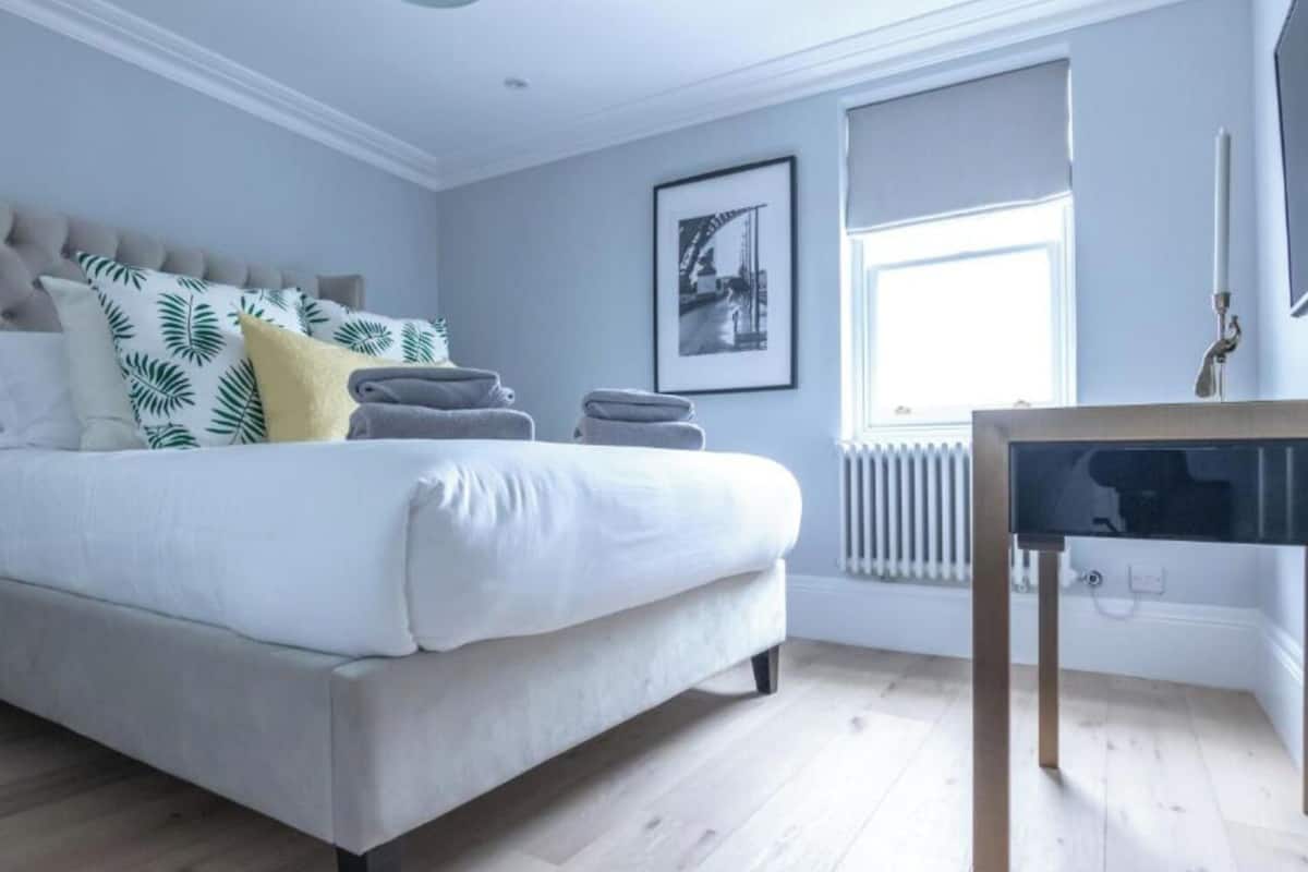 Een slaapkamer met een bed, kussens, een radiator, een raam en een tafel.