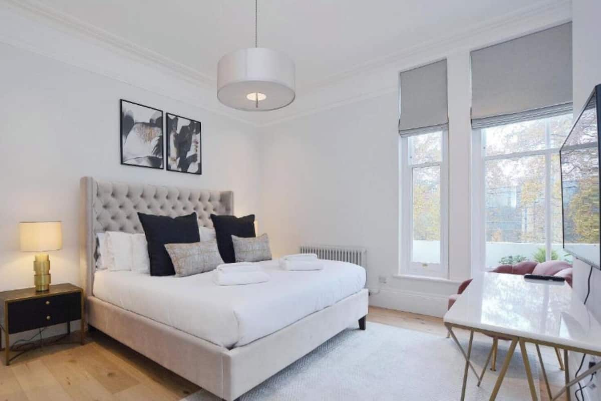 Een slaapkamer met een groot bed, een nachtkastje met lamp, een stoel, een bureau en een groot raam met jaloezieën.