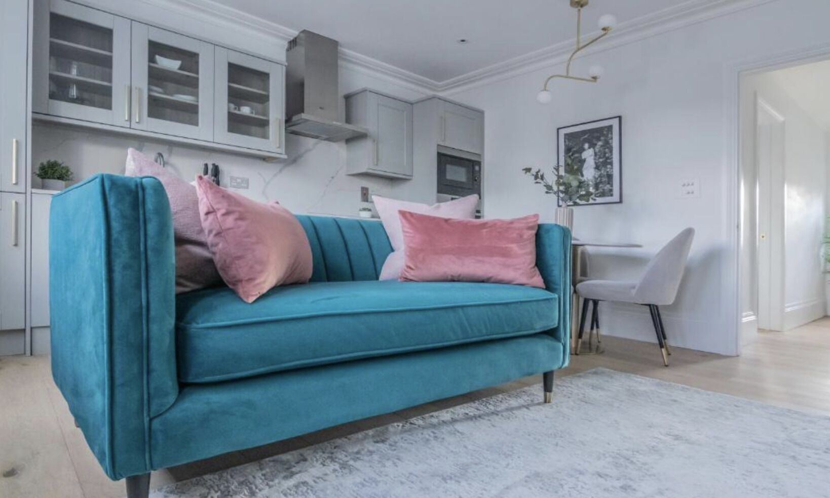 Un canapé turquoise agrémenté de coussins roses, dans un salon moderne avec une cuisine en arrière-plan.