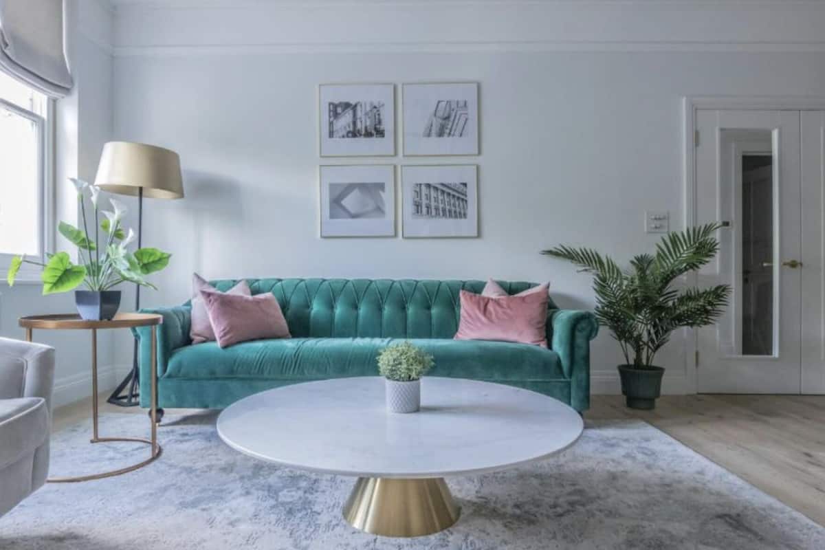 Een woonkamer met een turquoise bank, een ronde salontafel en een vloerlamp. Er hangen ingekaderde foto's aan de muur en er staat een potplant op de vloer.