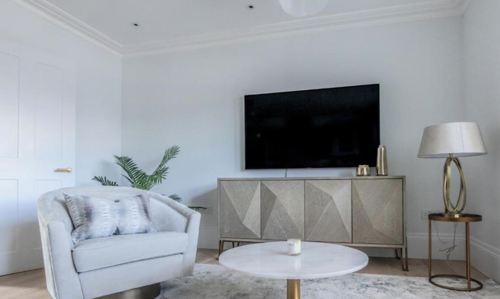 A modern living room with a white sofa, a round coffee table, a cabinet with geometric patterns, a flat-screen TV, and a floor lamp.
