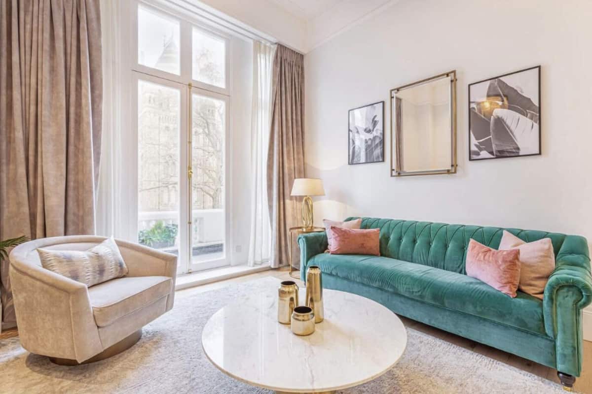 Een woonkamer met een beige fauteuil, een teal kleurige bank, een ronde salontafel en een groot raam met gordijnen.