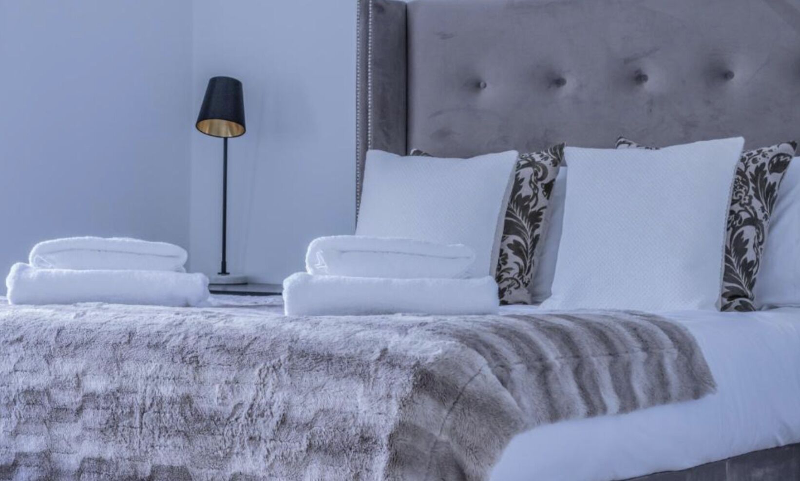 Een net opgemaakt bed met witte lakens en een grijze deken, een bedlampje en een gestoffeerd hoofdbord.