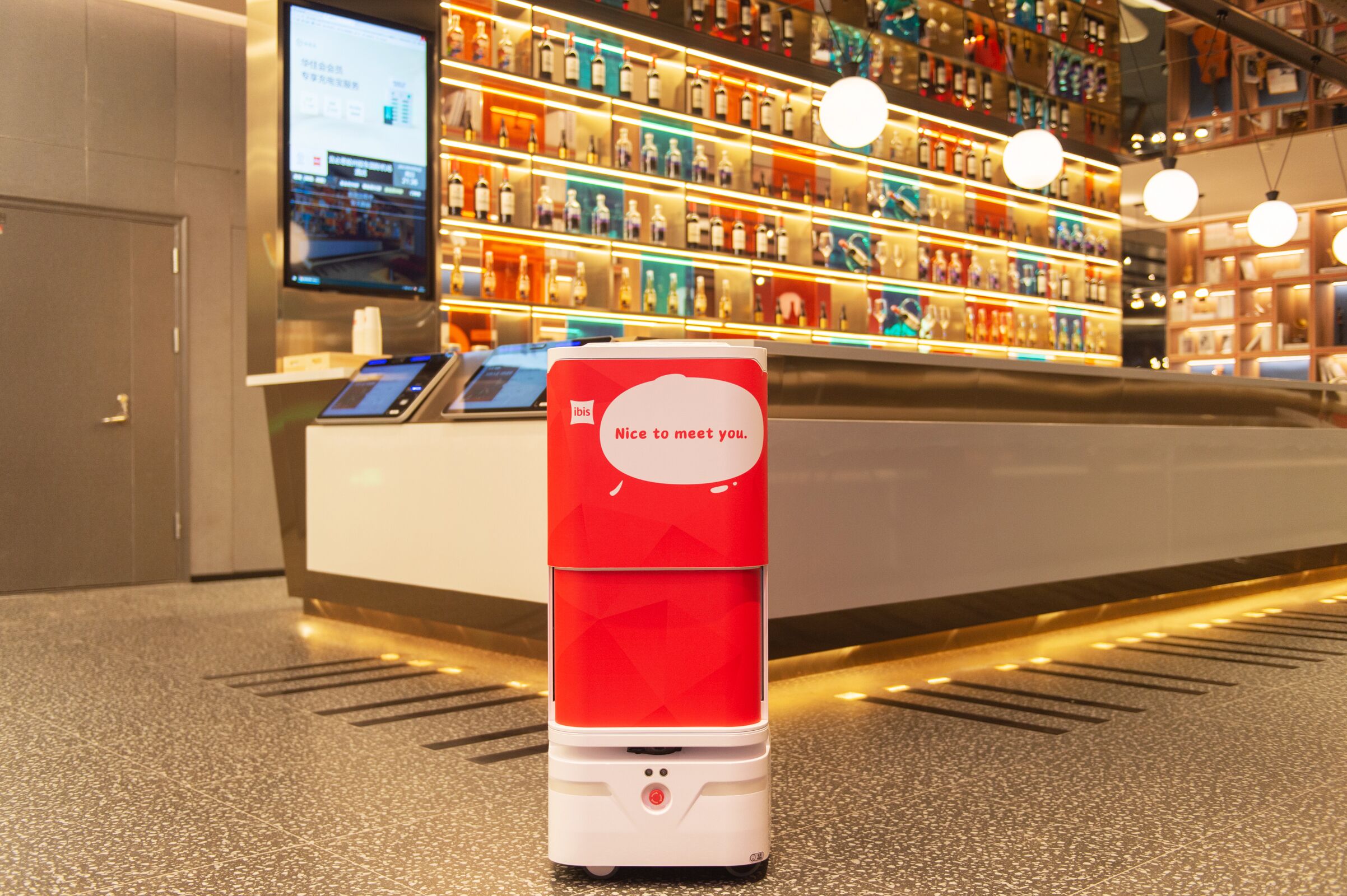 A red and white self-service kiosk in a hotel lobby with a digital screen and a message saying "Nice to meet you."