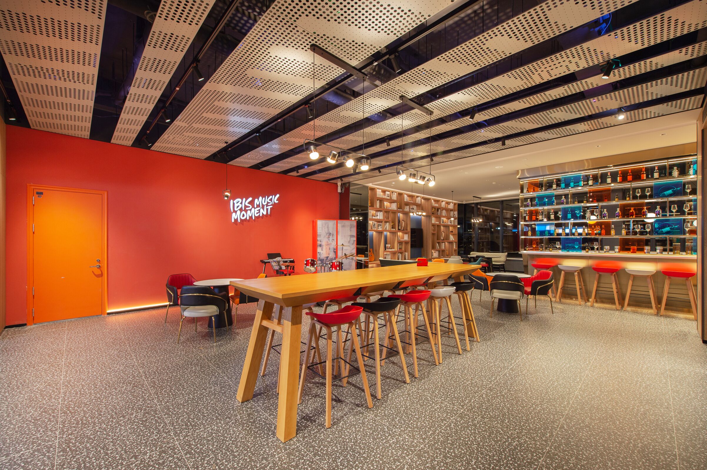 A modern interior with a long wooden table and red chairs, a bookshelf with various items, and a bright orange wall with the text "THIS MUSIC MOMENT".