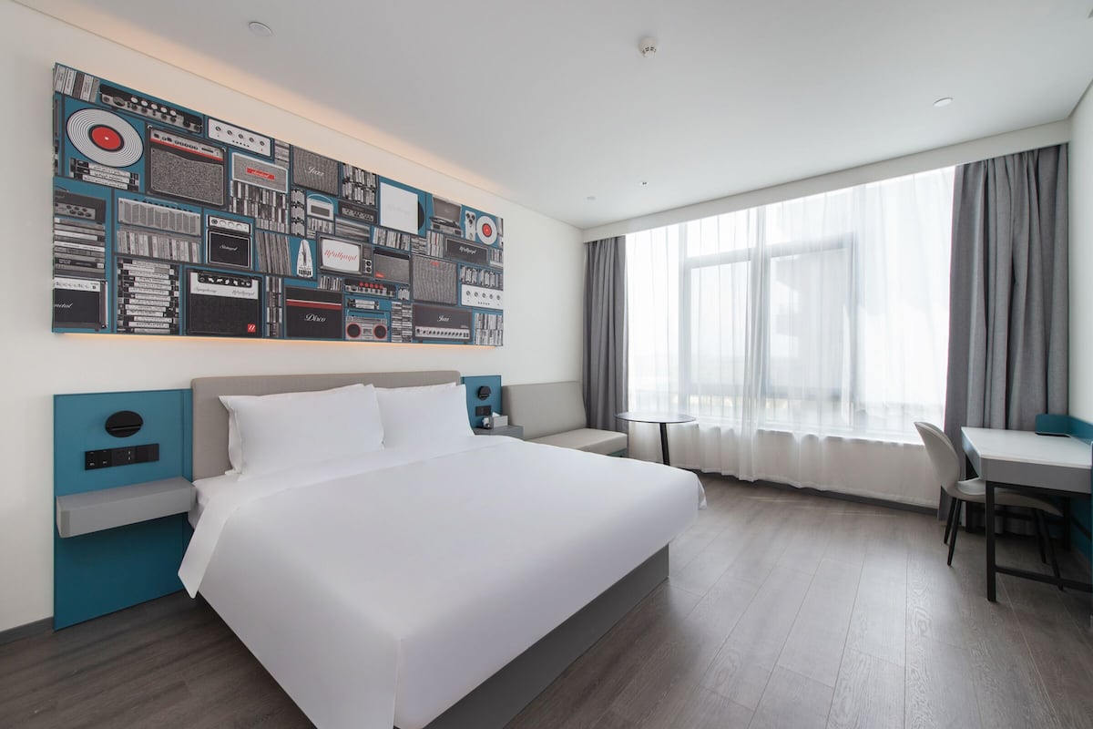 A modern hotel room with a large bed, a desk, a chair, a wall-mounted TV, and a large window with curtains.