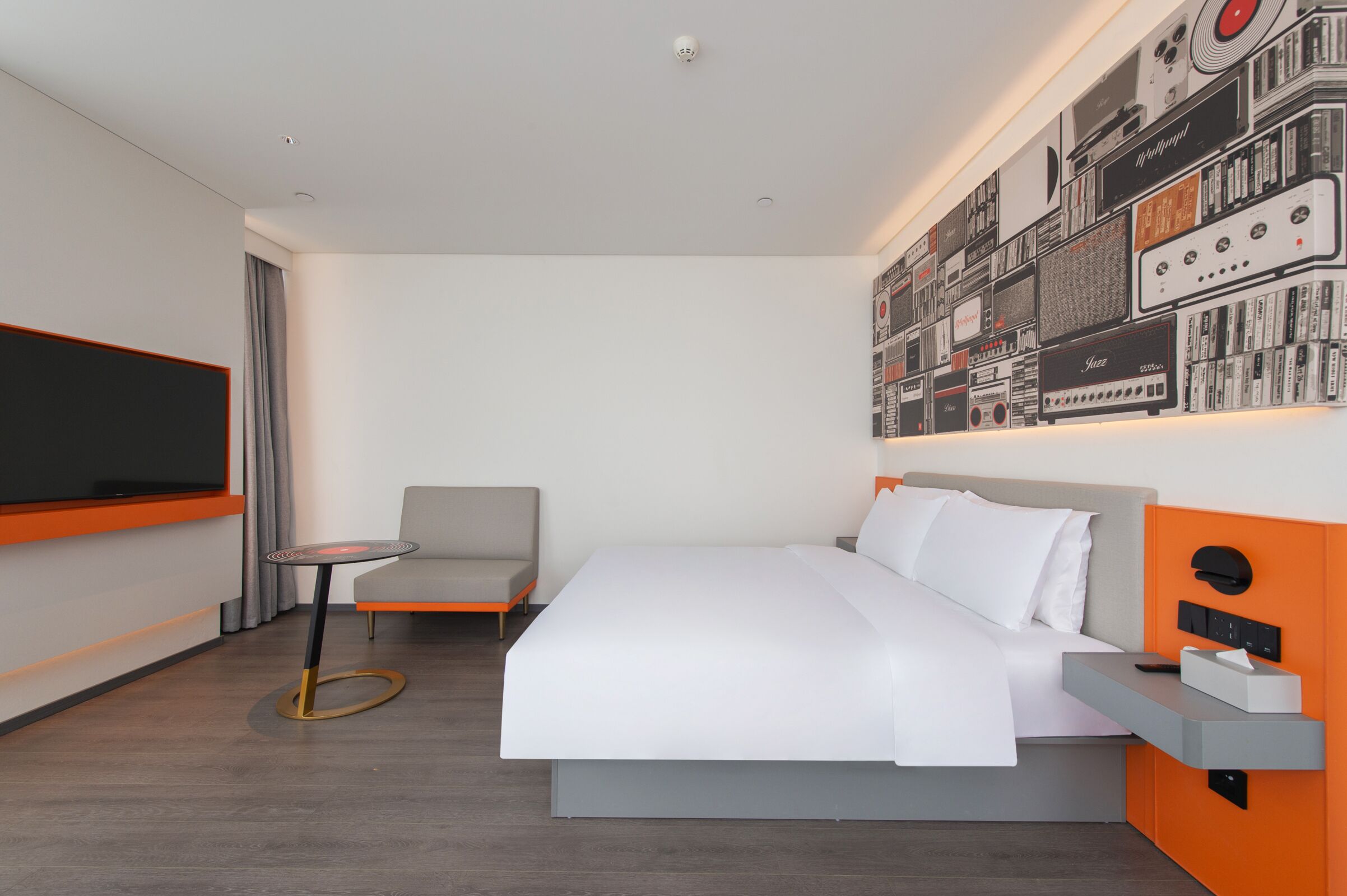 A modern hotel room with a large bed, a small table, a TV, and a wall decorated with vintage audio equipment.