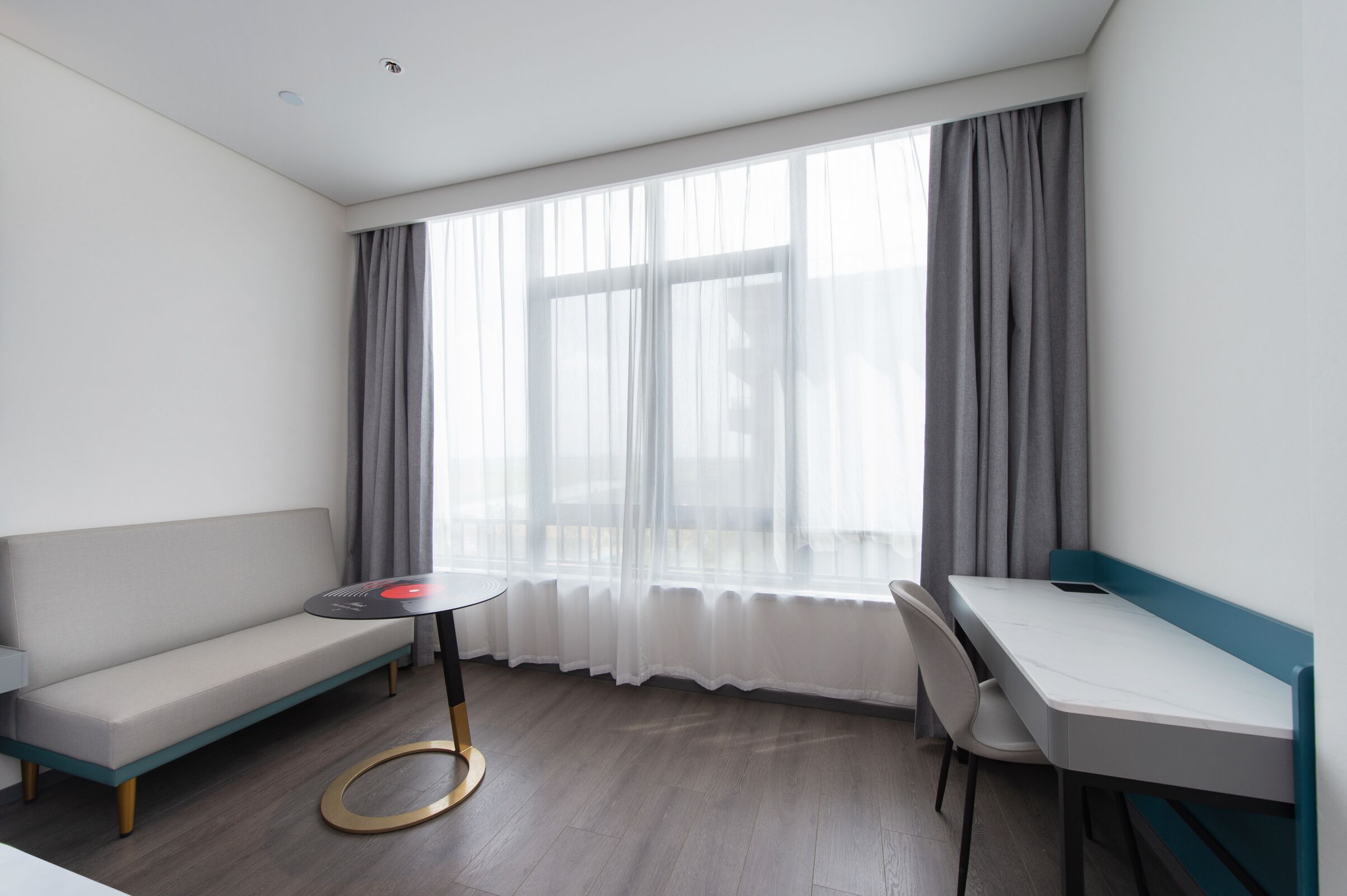 Una habitación moderna con sofá, una pequeña mesa redonda, escritorio y un amplio ventanal con cortinas translúcidas.