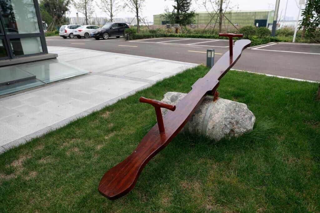 Una estructura de madera y metal, similar a un balancín, apoyada sobre una roca, en una zona con césped, un camino pavimentado y coches aparcados al fondo.