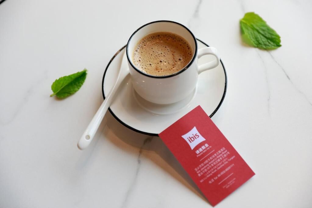 A cup of coffee on a saucer with a spoon, a red card, and a green leaf on a white surface.