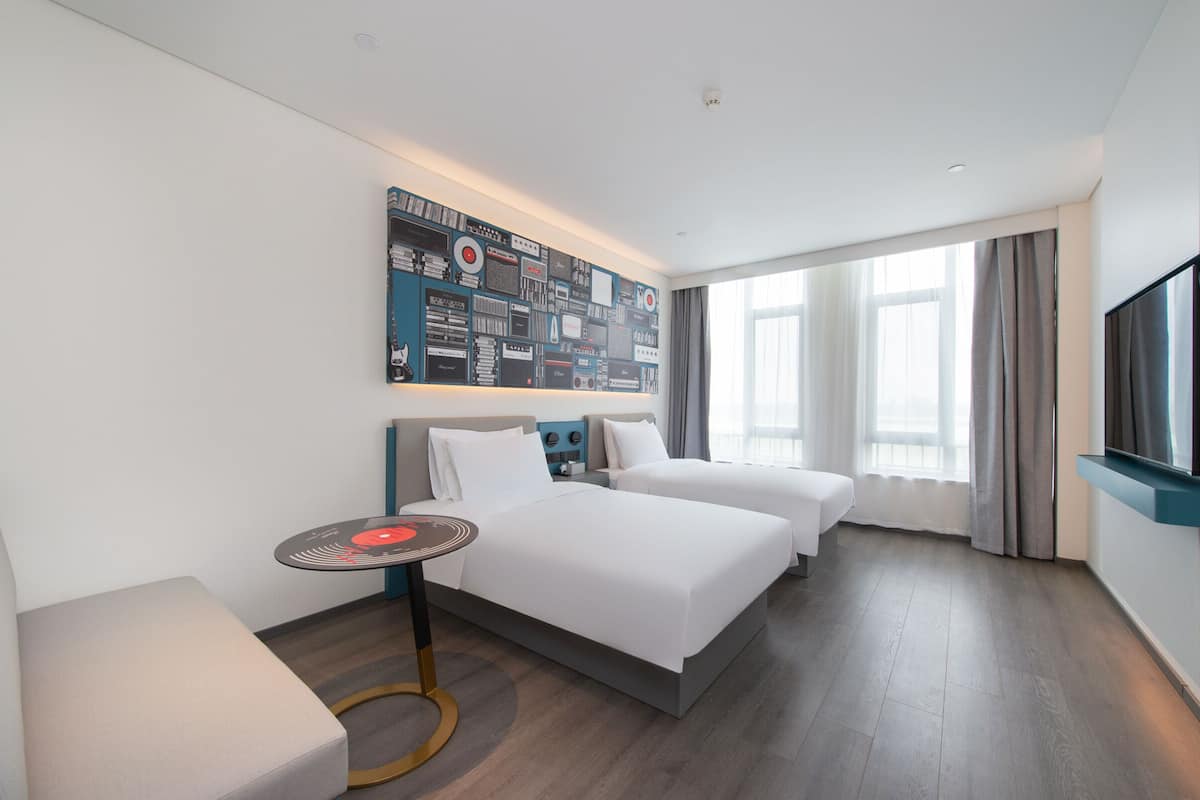 A hotel room with two beds, a small table, a wall-mounted TV, and a large window with curtains.