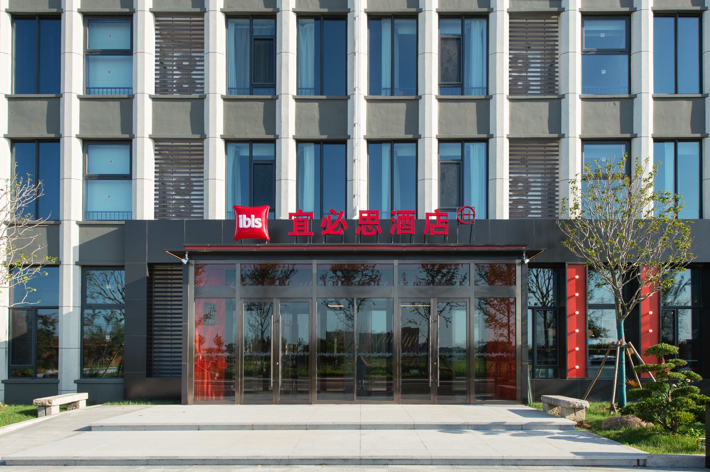 Une entrée d’hôtel moderne avec une façade en verre, une enseigne rouge et un petit arbre au premier plan.