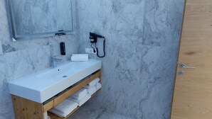 Un baño con lavamanos blanco, paredes de mármol, tocador de madera, toallero y una puerta.