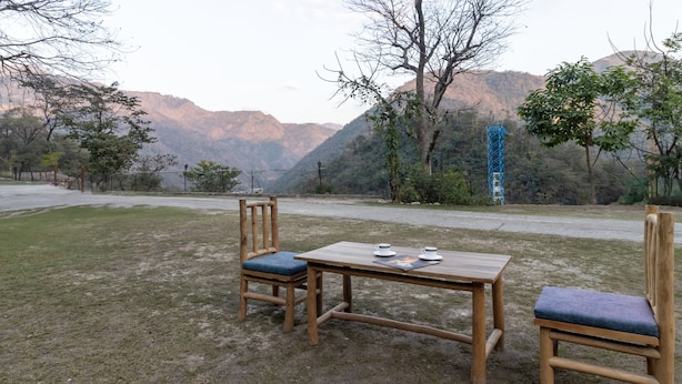 An outdoor seating area with a wooden table and chairs, set against a backdrop of mountains and a blue tower. - Itsy Hotels Paddler Zone With Bungee Jumping (Narendranagar)