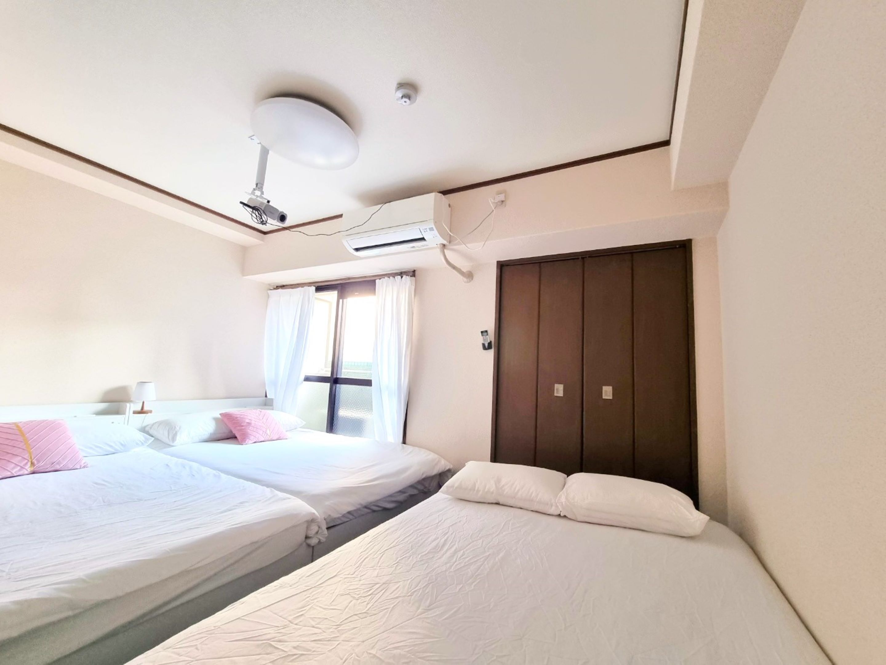 A hotel room with two beds, a ceiling fan, an air conditioning unit, a window with curtains, and a wardrobe.