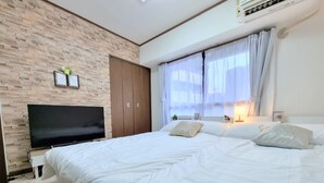 Une chambre avec un mur en briques, un téléviseur à écran plat, une armoire, un lit avec une literie blanche et une fenêtre ornée de rideaux légers.