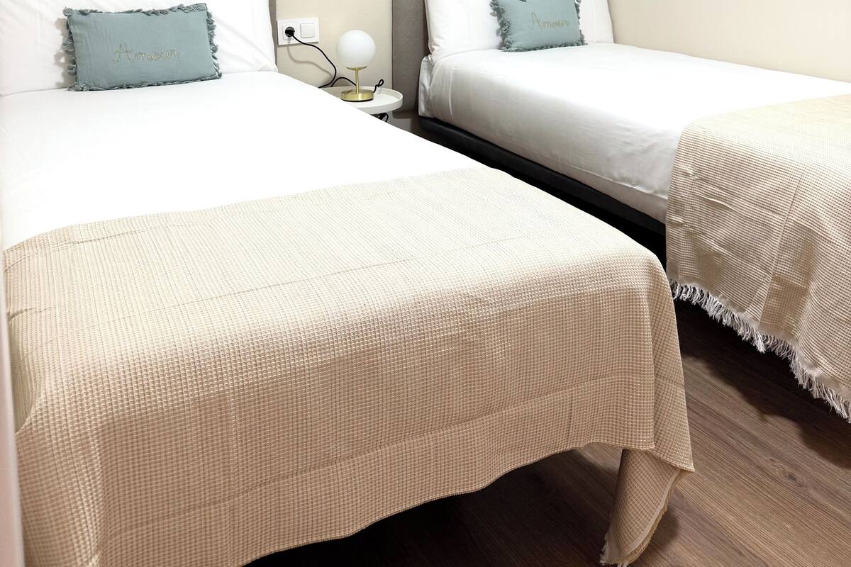 Ein Hotelzimmer mit zwei Betten, jedes mit einer weißen Bettdecke und einem blauen Kopfkissen. Ein Nachttisch mit Lampe und Steckdose ist vorhanden.